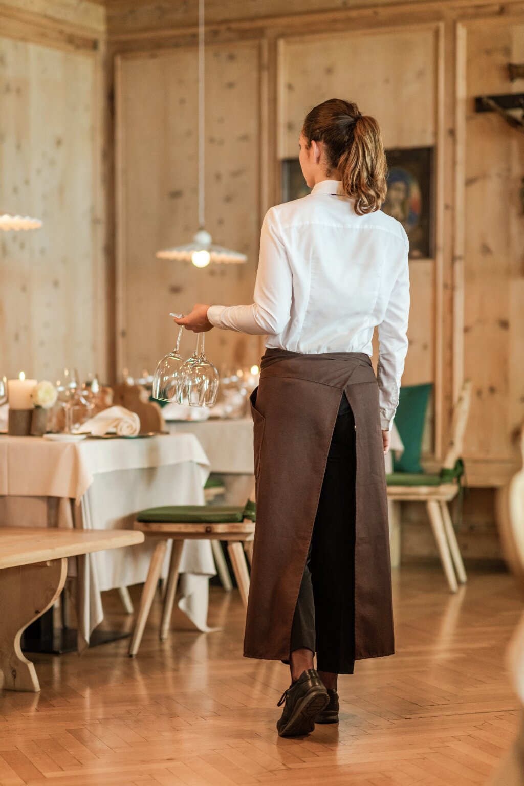 Foto: Armin Terzer Kellnerin mit langer Schürze in einem gedeckten Restaurant mit Holzvertäfelung.